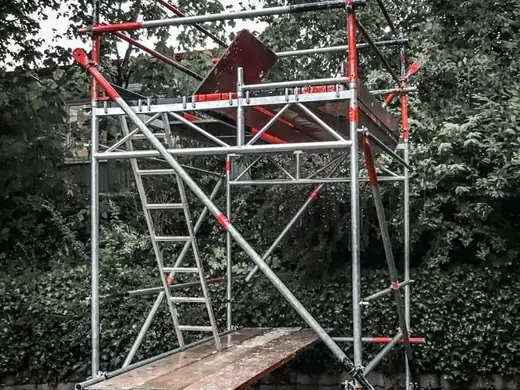 Auf einem Gehweg steht ein Gerüstturm mit einer Holzplattform und Treppen.